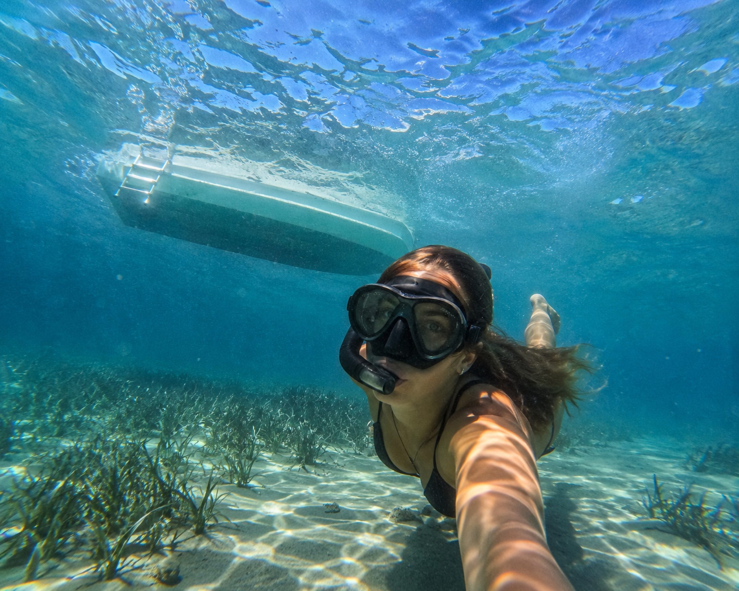 A snorkeller underwater over a sandy seabed with the hull of the boat above her
